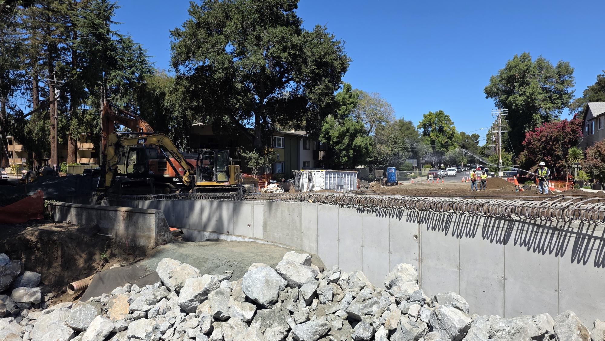 Bridge site showing excavators, concrete wall, rebar, and workers preparing the road