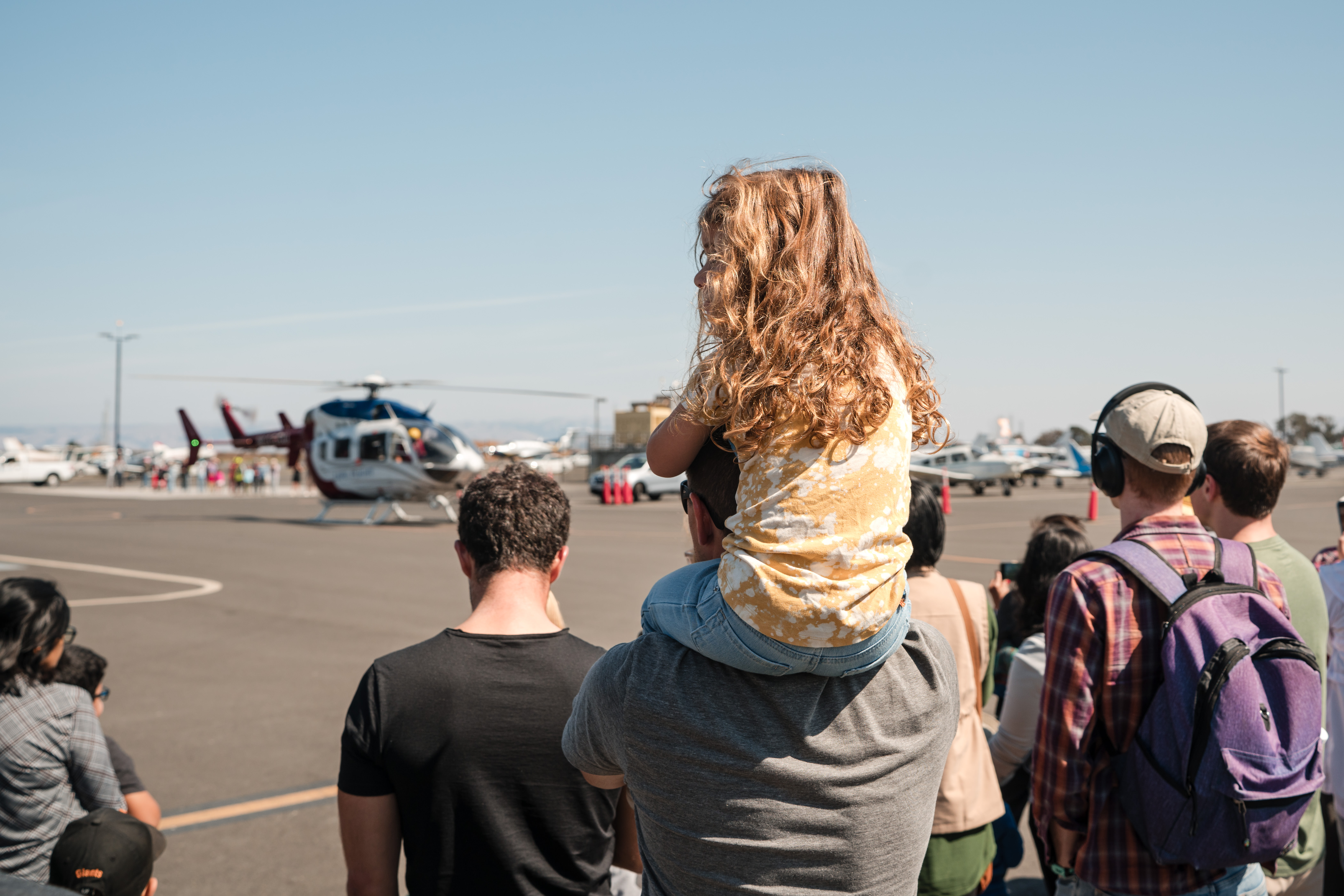 A girl sits on her dad's shoulders while they look at a helicopter on the tarmac