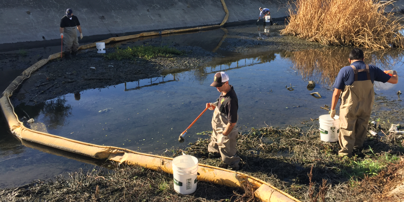 three people in waders use trash pickers and buckets to remove trash from behind a trash boom - a device similar to a giant pool noodle used to capture floating trash