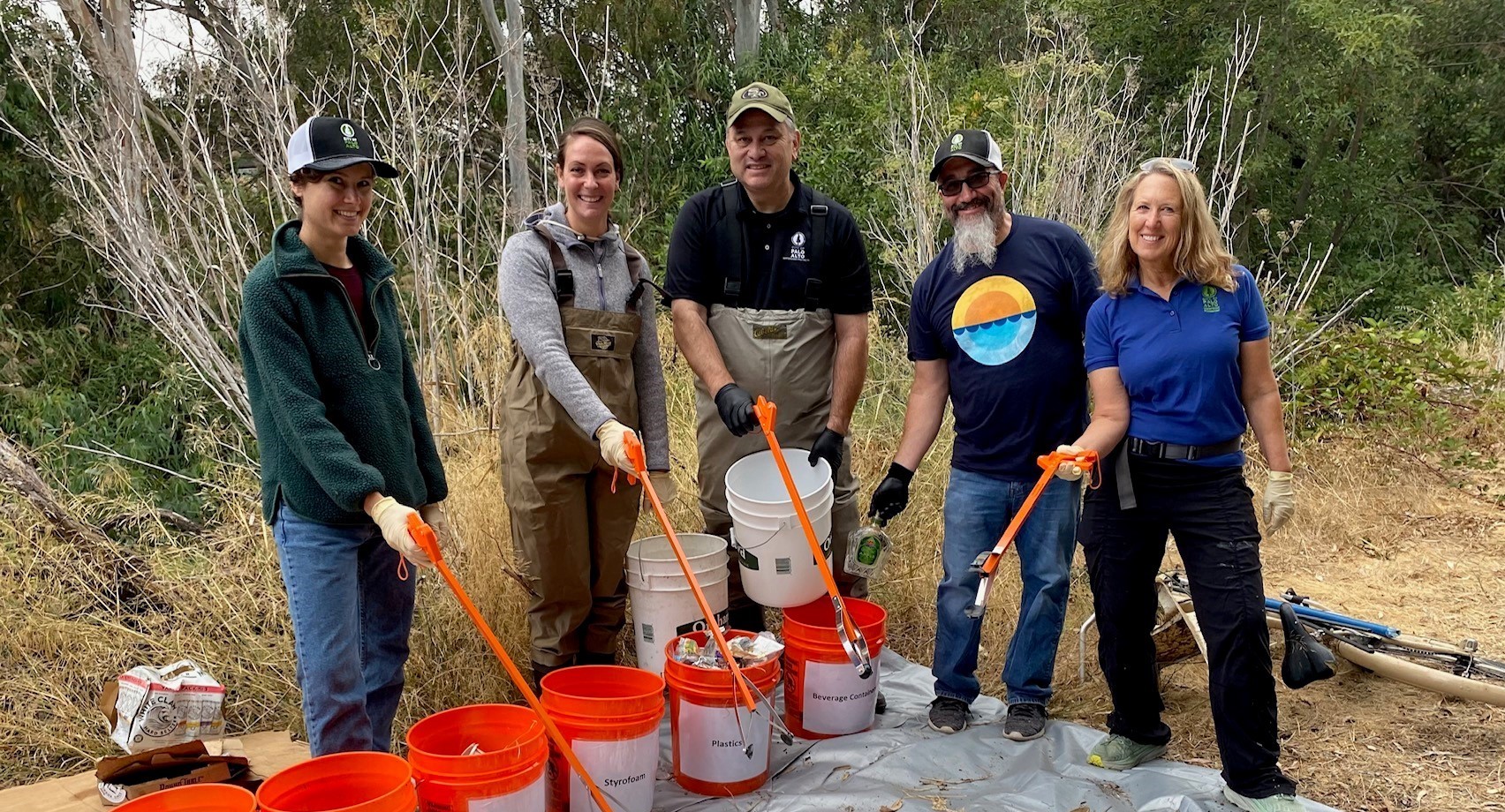 five people point bright orange trash pickers over a row of buckets at a small pile of trash on a tarp