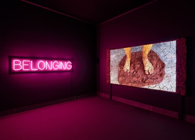 installation view of a neon sign that reads belonging, a clay wall with video projection